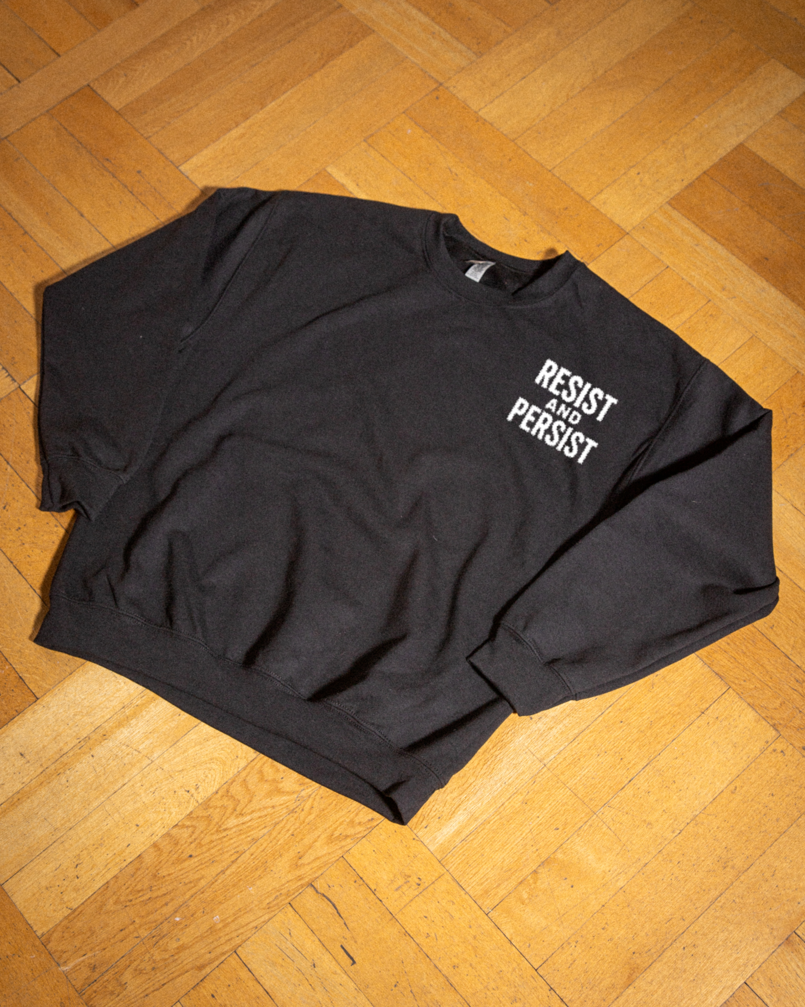 Resist & Persist Sweatshirt flat lay – black crewneck fleece with white chest graphic, Common Ground Project
