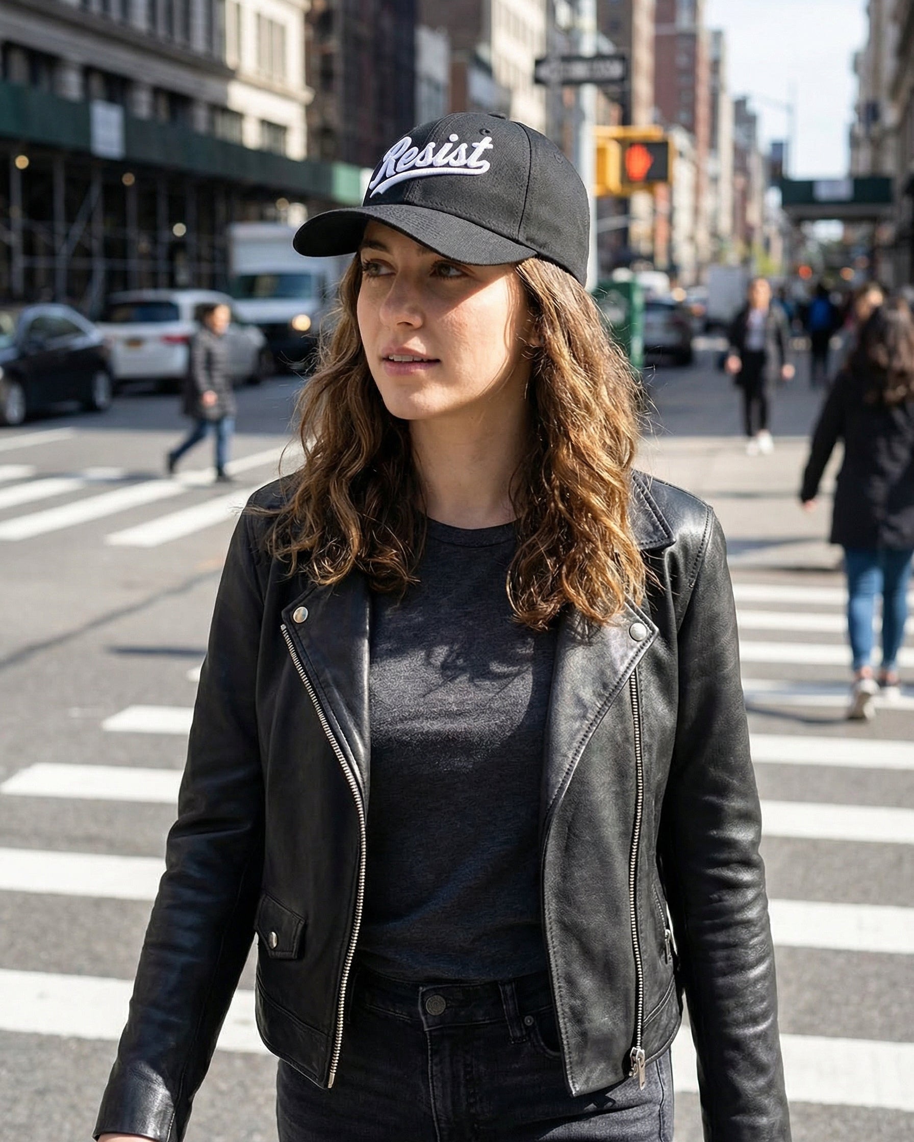 Person wearing a black leather jacket and 'Resist' cap on a city street.